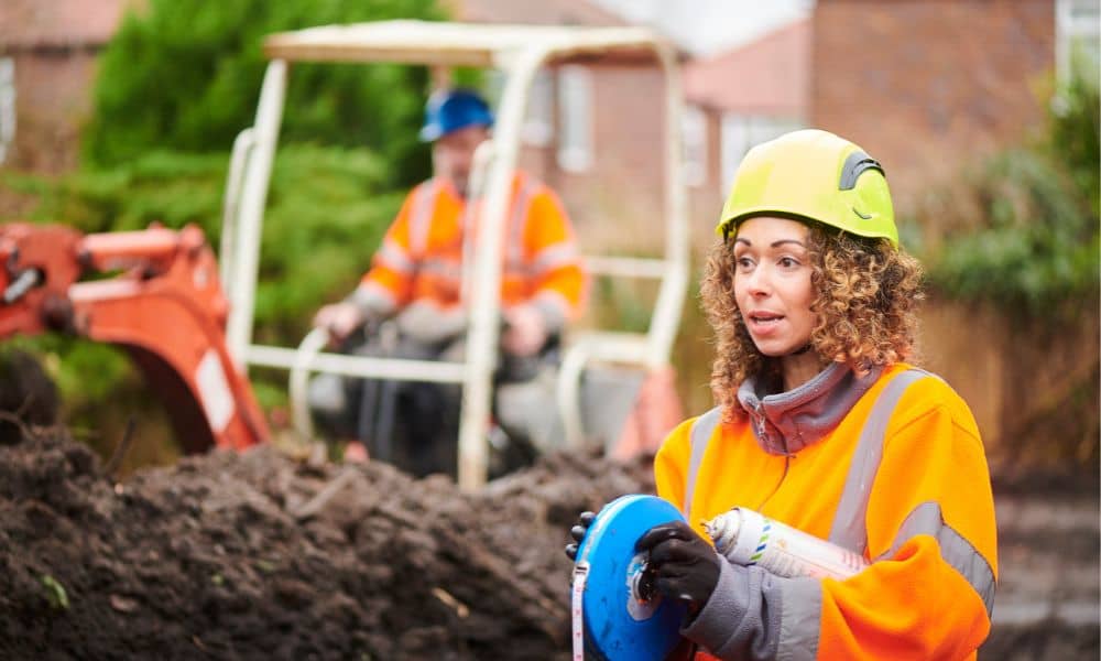 Female civil engineering professional in safety gear assessing a construction site with excavation equipment in the background
