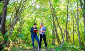 Two construction surveyors using a GNSS rover and field notebook under dense tree canopy showing RTK signal challenges